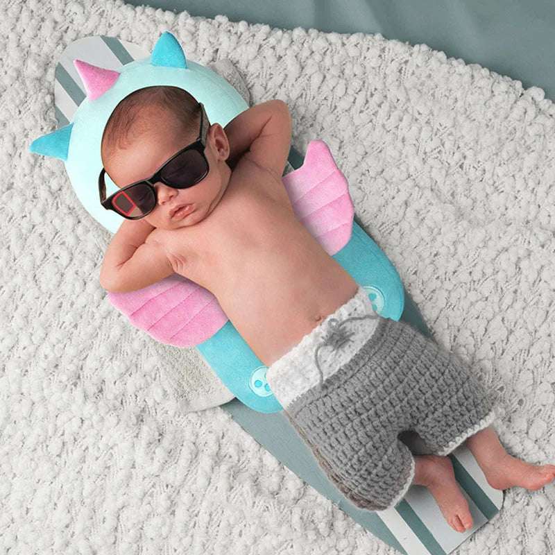 Baby wearing sunglasses and gray shorts lying on a plush dinosaur-themed cushion on a soft blanket