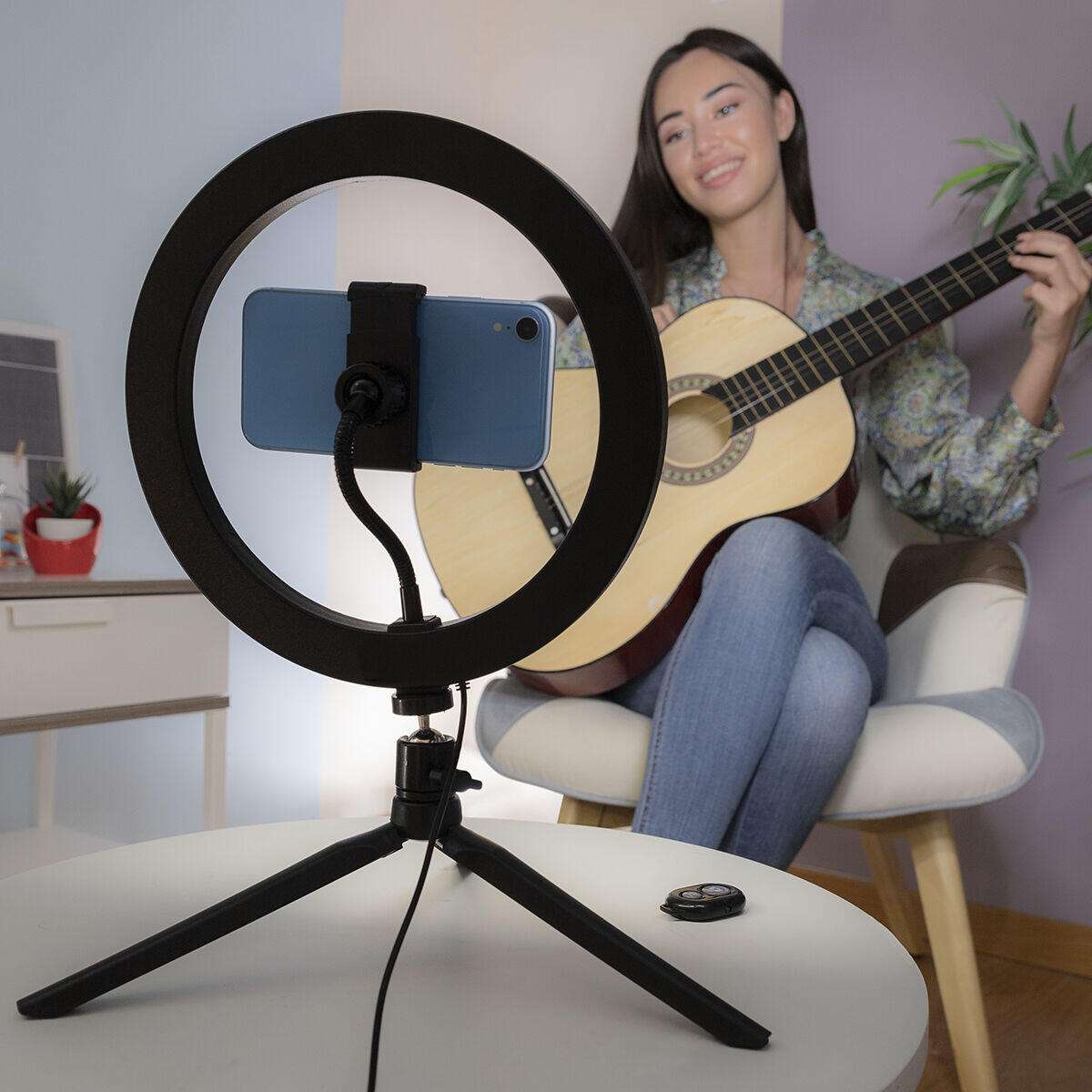 Ring light with phone holder on tripod, woman playing guitar in background, indoor recording setup
